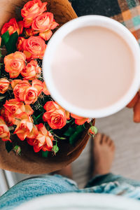 Woman holding white cup of coffee, latte cappucino, in background of bouquet orange roses flowers. 