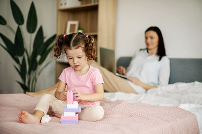 A cute girl is playing with a constructor on the bed while her mom is working on a laptop