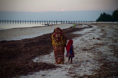 Rear view of mother and daughter holding hands while walking at beach against sky during sunset