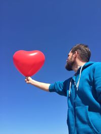 Low angle view of man holding heart shape balloon against clear blue sky