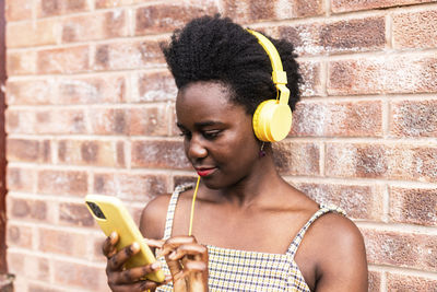 Young woman using smart phone and listening to music in front of brick wall