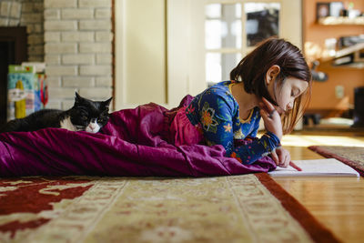 A child curls up on floor in a sleeping bag with cat doing schoolwork