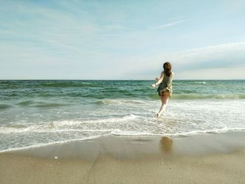 Rear view of happy woman running towards sea on shore at beach against sky