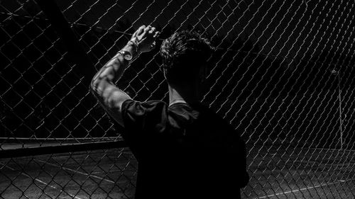 Rear view of man standing by chainlink fence at night