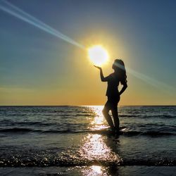 Optical illusion of silhouette woman holding sun at beach against bright sky during sunset