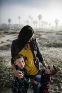 Cute sons embracing mother while standing at beach against sky during sunset