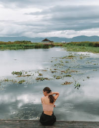 Rear view of woman sitting on pier over lake against sky