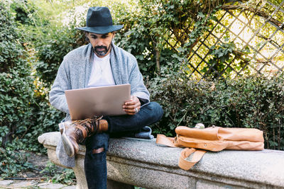 Adult hispanic man in trendy clothes and hat browsing netbook while sitting on stone bench and working remotely from park