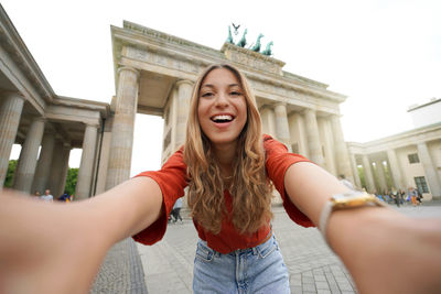 Woman takes selfie picture with smartphone camera in front of brandenburg gate, berlin, germany.
