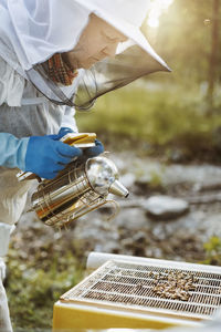 Mature beekeeper holding smoker while examining beehive on field