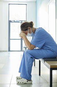 Exhausted nurse wearing protective face mask sitting on bench in corridor