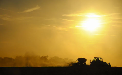 Silhouette of a tractor sowing seeds in a field in a cloud of dust against the background.