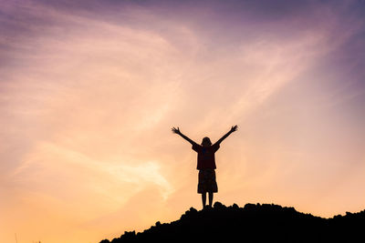 Silhouette boy with arms raised standing against sky during sunset