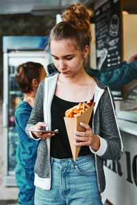 Teenage girl eating potato fries from carton cone standing in front of food truck