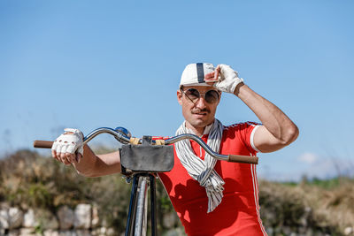 Stylish adult male with mustache in sunglasses and bright red shirt in composition with scarf and gloves touching striped cap while leaning on bike handlebar against blurred grassed hill and blue sky in nature
