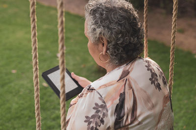 Back view of senior lady wearing blouse sitting in park and reading electronic book