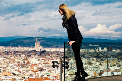 Young woman wearing warm clothing while standing on balcony against cityscape