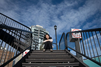 Low angle portrait of smiling young woman sitting on steps against sky
