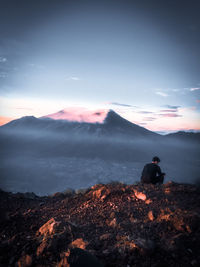 Rear view of woman sitting on mountain against sky during sunset