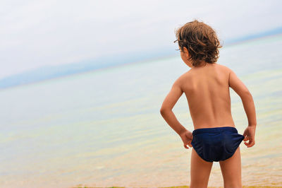 Rear view of shirtless boy standing at beach against sky