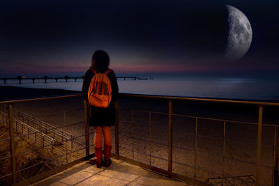 Rear view of woman with backpack looking at sea against sky during night