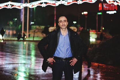 Portrait of man with hands on hip standing on footpath at night