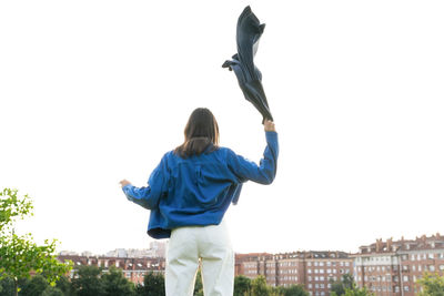 Back view of anonymous female in trendy wear waving scarf on background of sky in city on windy day