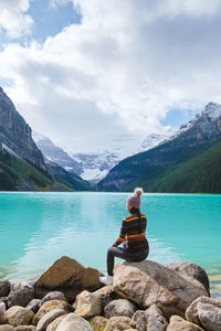 Rear view of woman sitting on rock by lake against sky