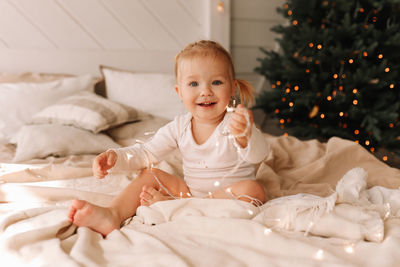 Happy cheerful baby little girl sitting smiling on a cozy bed at christmas holidays at home