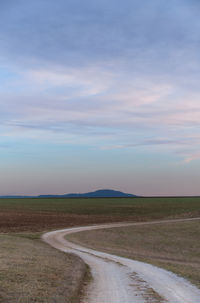 Dirt road passing through field against sky