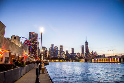 Illuminated buildings by river against clear sky