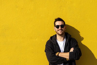 Young bearded man standing against a yellow wall with crossed arms