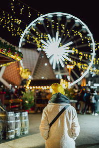 Rear view of woman standing against illuminated string lights at night