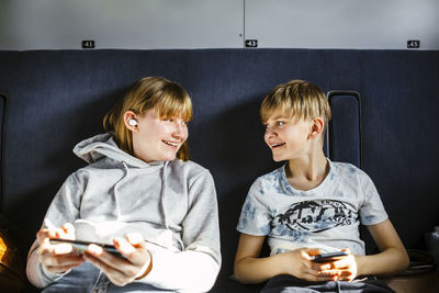 Smiling brother and sister with smart phones looking at each other while traveling in train