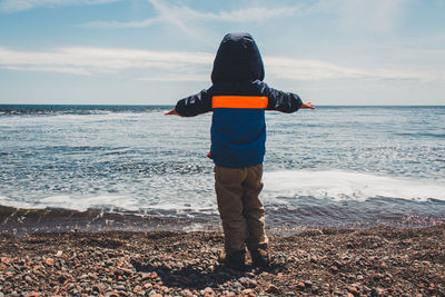 Rear view of boy in hooded jacket standing on shore against sky