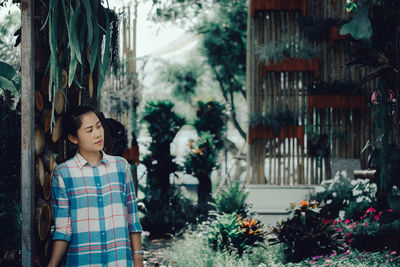 Woman looking away while standing by wall against plants in greenhouse