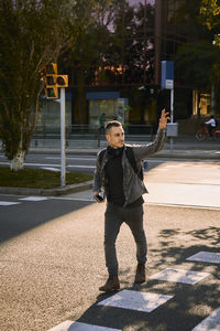 Young male crossing road in city and waving hand while looking away and catching taxi