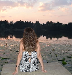 Rear view of woman sitting on pier over lake against sky during sunset