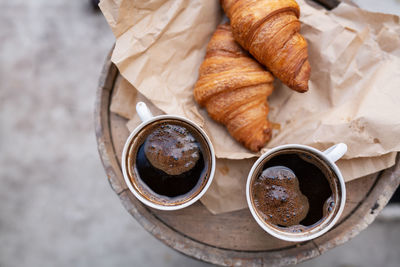 There are two cups of coffee and croissants on craft paper on a wooden barrel. retro style
