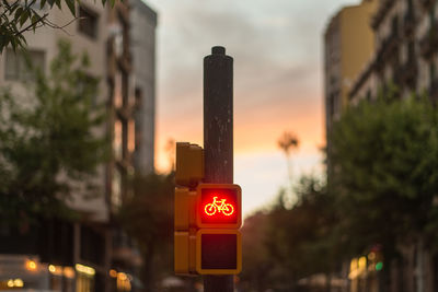 Bicycle sign on pole in city against sky during sunset