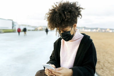 Woman with curly hair wearing face mask using smart phone while sitting against clear sky