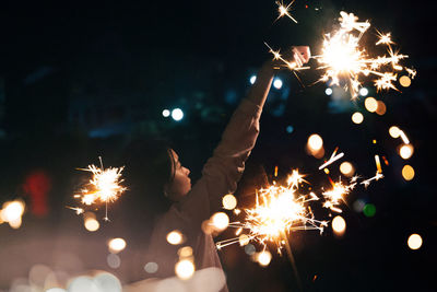 Young woman holding burning sparkler while standing outdoors at night