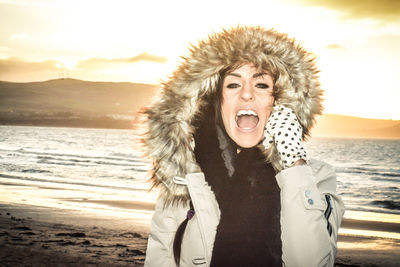 Portrait of woman shouting while wearing fur coat against sea at beach
