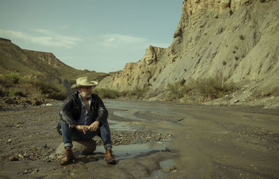 Adult man in cowboy hat sitting on rock along river flows in desert. almeria, spain