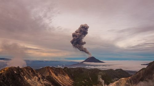 Smoke emitting from mountain against cloudy sky