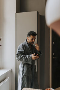 Man in bathrobe using smart phone while brushing teeth at home