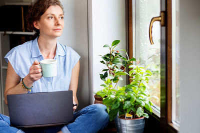 Thoughtful woman holding coffee looking through window at home