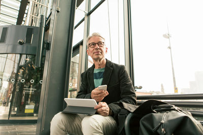 Low angle view of senior male commuter looking away sitting with technologies by bags at railroad station