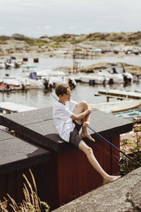 Smiling boy with fishing rod sitting on rooftop at harbor during vacation