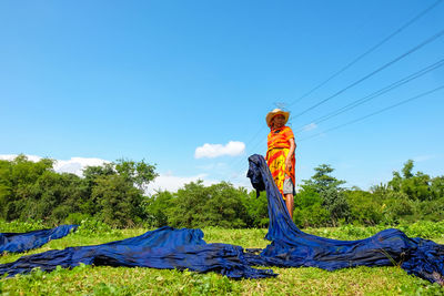 Mature woman drying blue fabrics on grassy field against blue sky during sunny day